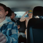 Image of the inside view of a car with a young male driver sitting behind the steering wheel, with both hands on the wheel. Two of his friends are sat in the back of the car. One is a young woman wearing rugby kit, and the other is a young man wearing swimming trunks, a swimming cap, and goggles.