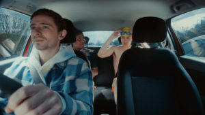 Image of the inside view of a car with a young male driver sitting behind the steering wheel, with both hands on the wheel. Two of his friends are sat in the back of the car. One is a young woman wearing rugby kit, and the other is a young man wearing swimming trunks, a swimming cap, and goggles.