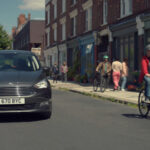 A man cycling, being passed wide and slow by a car