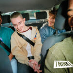 A photo of three young men in the back of a car wearing seatbelts, shot from the front.